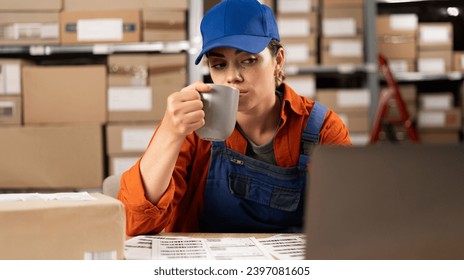 Warehouse Manager drinking tea or coffee working in storeroom with rows of shelves of parcels, packages ready for shipment. Break on work. Copy space - Powered by Shutterstock