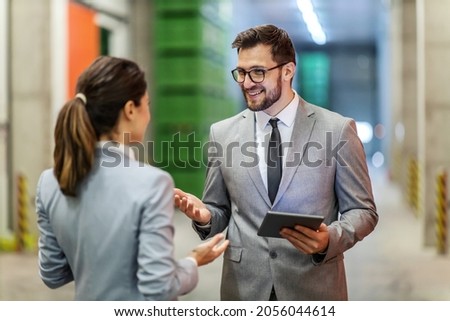 Warehouse manager training. A man and a woman in a business suit stand in a warehouse and talk about work. A smiling man with glasses holds a tablet in his hand and looks the woman straight in the eye