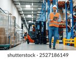 Warehouse manager using digital tablet is walking through warehouse and checking inventory, in background forklift is passing by