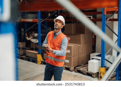 Warehouse manager wearing high visibility vest and hardhat using digital tablet, managing logistics and doing inventory in a large distribution center - Powered by Shutterstock