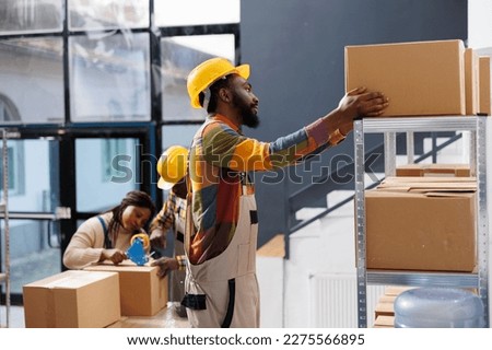 Warehouse package handler choosing cardboard box on shelf while standing on ladder. African american man storehouse worker preparing customer parcel and taking carton container
