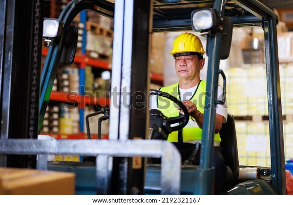 Warehouse staff driving forklift operator\
moving boxes in industrial container warehouse factory. Asian\
senior Man worker woking with\
forklift.
