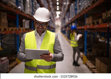 Warehouse Supervisor Reading Report On Tablet About Successful Delivery And Distribution In Warehouse Logistics Center. In Background Coworker Checking Inventory And Productivity In Storage Area.