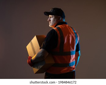 Warehouse Worker With Cardboard Box. Warehouse Worker Male. Portrait Of A Warehouse Fulfillment Employee On A Dark Background. He Is Thinking About Something. Fulfillment Center Employee.