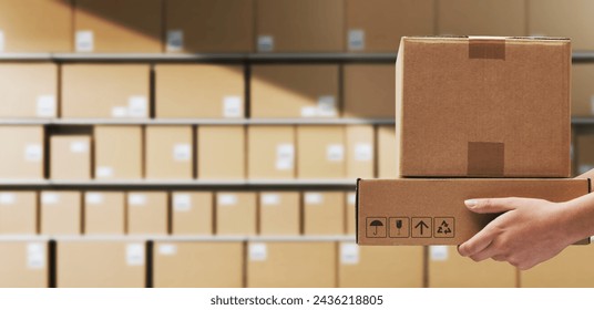 Warehouse worker holding delivery boxes and stacked packages in the background, logistics and shipment concept - Powered by Shutterstock