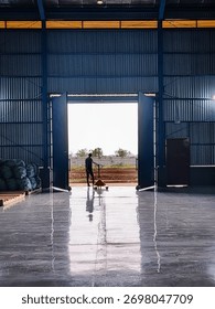 A warehouse worker pulling a hand pallet jack through the open gate of an industrial factory building. The man stands in the center of the warehouse entrance, looking outside toward the bright sunligh