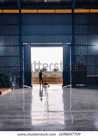 A warehouse worker pulling a hand pallet jack through the open gate of an industrial factory building. The man stands in the center of the warehouse entrance, looking outside toward the bright sunligh