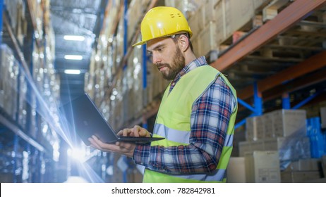 Warehouse Worker Uses Laptop. He's Standing In The Middle Of  A Big Distribution Center With Big Storage Racks And Pallets On Them.