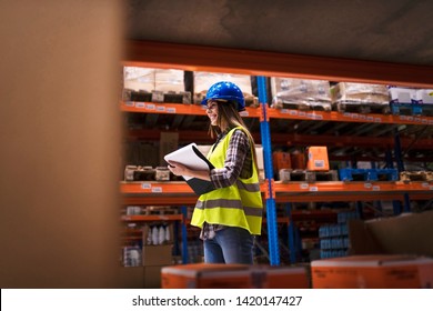 Warehouse Worker Walking Through Large Storage Area Checking Inventory And Organizing Goods Distribution. Smiling Woman Employee Working In Warehouse Distribution Center.