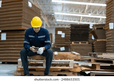 Warehouse worker wearing a yellow hard hat and gloves takes a break to check his phone. Stacks of cardboard sheets are neatly arranged on wooden pallets in the background. - Powered by Shutterstock