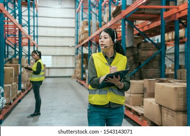 Warehouse worker woman wearing headset holding clipboard in large warehouse. blurred view colleague in safety vest standing stock taking. lady staff answering phone call and talking by microphone - Powered by Shutterstock