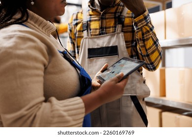 Warehouse workers checking inventory data statistics on tablet screen. African american logistics and distribution managers analyzing goods purchase analytics report in storehouse
