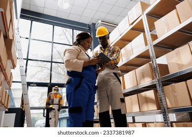 Warehouse workers looking at inventory data on digital tablet screen while standing in stockroom. African american logistics manager showing colleague goods storage management application