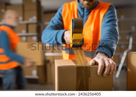 Warehouse workers preparing delivery boxes for shipping, an employee is sealing a box using a tape dispenser gun
