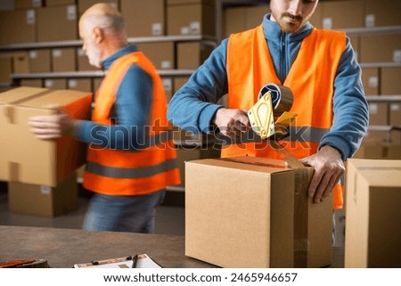 Warehouse workers preparing delivery boxes for shipping, an employee is sealing a box using a tape dispenser gun