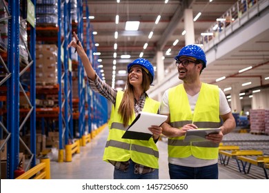 Warehouse Workers In Protective Uniform Walking Through Large Distribution Center Organizing Goods Distribution. Woman Employee Pointing To The Shelves While Her Coworker Using Tablet.