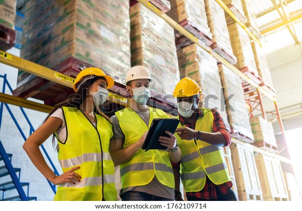 Warehouse\
workers team meeting using tablet computer with wearing face mask\
and protective safety helmet standing in storage factory.standard\
to prevent coronavirus COVID-19\
infection