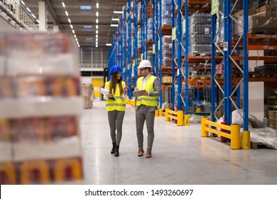 Warehouse Workers Walking Through Large Storage Department Discussing About Distribution And Logistics. Warehouse Interior With Palette Of Goods And Shelves.