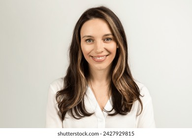 A warm and approachable european young woman with long brown hair and a welcoming smile poses in a white blouse, standing against a soft neutral background, studio. Work, business meeting - Powered by Shutterstock