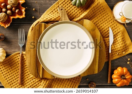 Warm autumn table setting with white plate on cutting board, yellow cloth, rustic cutlery, small pumpkin, pinecones, nuts, and ceramic candle. Cozy flat lay ideal for Thanksgiving