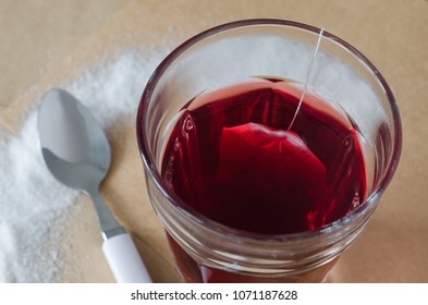 Warm Berry Tea And Tea Bag In Glass, Spilled Sugar Around And White Spoon Near, On Bright Brown Paper Background