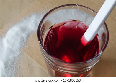 Warm Berry Tea And Tea Bag In Glass, Spilled Sugar Around And White Spoon In Tea, On Bright Brown Paper Background