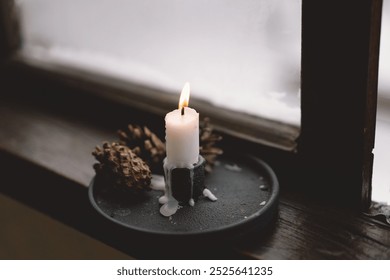 A warm candle flame flickers against a soft backdrop while resting on a dark holder next to pine cones, creating a cozy atmosphere in the room. - Powered by Shutterstock