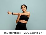 Warm up concept. Young slim african american woman stretching her arms before training, standing over blue background and looking at camera, wearing activity tracker