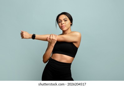 Warm up concept. Young slim african american woman stretching her arms before training, standing over blue background and looking at camera, wearing activity tracker