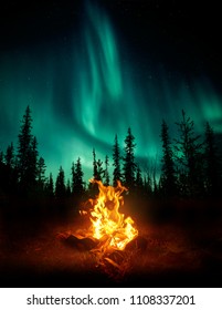 A Warm And Cosy Campfire In The Wilderness With Forest Trees Silhouetted In The Background And The Stars And Northern Lights (Aurora Borealis) Lighting Up The Night Sky. Photo Composite.