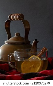 Warm And Cozy Studio Shot Of A Cinnamon Hot Toddy Alcoholic Drink In A Clear Mug, With Cinnamon Stick And Lemon Wheel Nestled In Black And Red Flannel With A Vintage Copper Kettle In The Background.