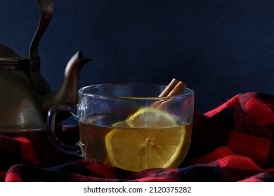 Warm And Cozy Studio Shot Of A Cinnamon Hot Toddy Alcoholic Drink In A Clear Mug, With Cinnamon Stick And Lemon Wheel Nestled In Black And Red Flannel With A Vintage Copper Kettle In The Background.