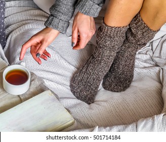 Warm And Cozy,and Warm Concept , Girl Sitting On White Bed In A Sweater And Knitted Socks With A Book And A Cup Of Tea.On The Table A Plate Of Cookies, Flowers And Candle.