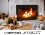 Warm cup of tea, pumpkin, autumn leaves on wooden table on background of burning fireplace. Cozy autumn still life. Thanksgiving and Halloween. Fall hygge