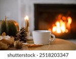 Warm cup of tea, pumpkin, autumn leaves on wooden table on background of burning fireplace. Cozy autumn still life. Thanksgiving and Halloween. Fall hygge