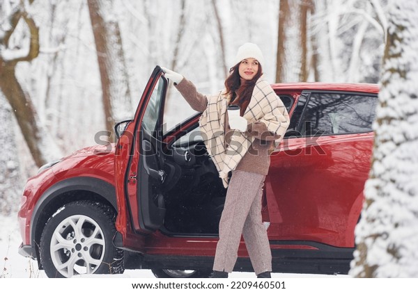 Warm drink in cup. Beautiful\
young woman is outdoors near her red automobile at winter\
time.