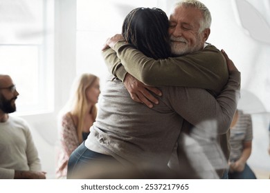 A warm embrace between two people in a group setting. Others are seated in the background, creating a supportive atmosphere. Smiles and hugs convey warmth and connection. Supportive group with elderly - Powered by Shutterstock