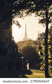 Warm evening light highlights a distant spire above rooftops, with leafy trees and dark stone buildings creating a natural frame around the urban scene