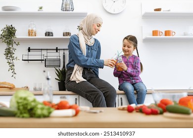 Warm family moment focusing on health and bonding. Muslim mother and daughter enjoying healthy juice together in modern kitchen. They are sitting happily amidst fresh vegetables and cozy surroundings - Powered by Shutterstock