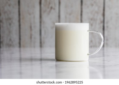 Warm frothy milk in a transparent glass cup. The cup is on a white reflective surface with a rustic white  wooden background. - Powered by Shutterstock