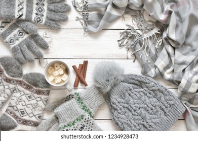 Warm grey woolen knitwear: socks, gloves, pom hat, scarf and cup of cocoa with marshmallow on white rustic wood background. Winter cozy still life. 
