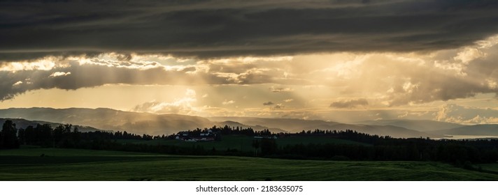 Warm, Hazy Sunshine And Sun Rays Come Through Dark Clouds Over A Shaded Misty Landscape With A Farm, Fields And Trees In The Distance. Ringsaker, Norway, June 28. 2022