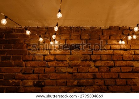 Warm incandescent string lights draped along a ceiling cast a soft amber glow over a textured exposed brick wall. The scene shows weathered red-brown bricks