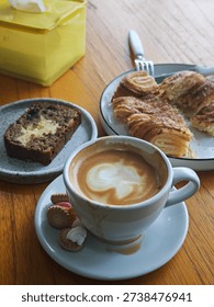 A warm and inviting breakfast scene is captured on a wooden table, centered around a cup of freshly brewed cappuccino topped with soft, creamy milk foam forming a delicate pattern on the surface. The coffee is served in a white ceramic cup placed on a matching saucer, accompanied by small assorted cookies that add a touch of sweetness and color.

Beside the coffee, a golden-brown croissant rests on a white plate, its flaky layers visibly crisp and buttery, with a portion already torn open to reveal its soft interior. A fork and knife lie nearby, suggesting a relaxed and unhurried meal. To the left, a slice of moist banana bread sits on a small ceramic plate, lightly spread with butter that melts gently into the bread’s textured surface.

In the background, a bright yellow tissue box adds a subtle pop of color, balancing the warm tones of the wooden table and the pastries. Soft natural light illuminates the scene, highlighting the textures of the coffee foam, the croissant’s layers, and the rustic table surface. Overall, the image conveys a cozy, calm atmosphere, evoking the simple pleasure of enjoying a leisurely morning coffee and breakfast.