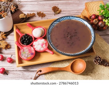 A warm and inviting dessert setup featuring a bowl of sweet brown soup with various colorful toppings, including boba and jelly, arranged on a wooden tray surrounded by autumnal elements