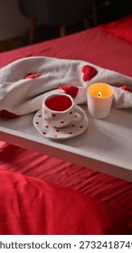 A warm and inviting scene featuring a heart-patterned teacup and saucer with red tea, a lit candle, and a soft blanket on a bed adorned with red sheets