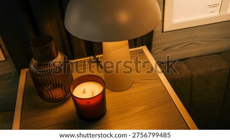 A warm and inviting scene featuring a lit candle in a red glass holder, a ribbed amber glass vase, and a contemporary mushroom-shaped lamp on a wooden nightstand