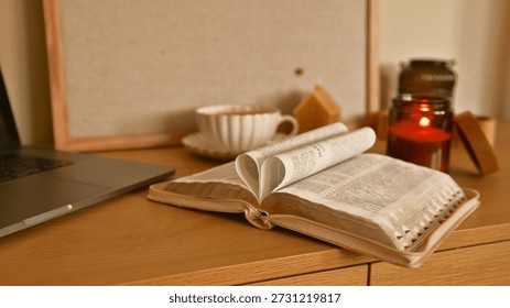 A warm and inviting scene featuring an open book with pages folded into a heart, alongside a laptop, coffee cup, and lit candle on a wooden desk