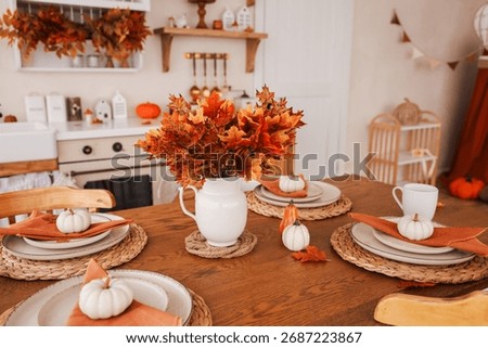 A warm kitchen features a dining table set for a meal decorated with orange leaves small pumpkins and napkins. The cozy atmosphere captures the essence of autumn festivities and gatherings.