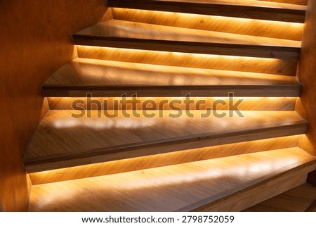 Warm light glows under each step. Wooden stairs curve gently upward. Soft illumination enhances texture and depth. Modern design blends comfort with elegance. Ideal for luxury interiors, upscale homes